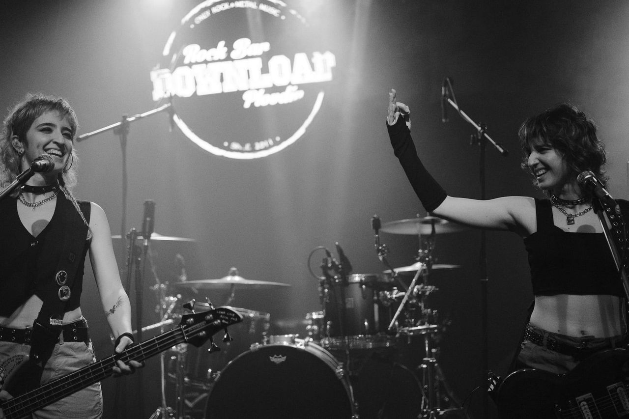 Hipersona’s two musicians smiling with guitars on a black-and-white indoor stage.