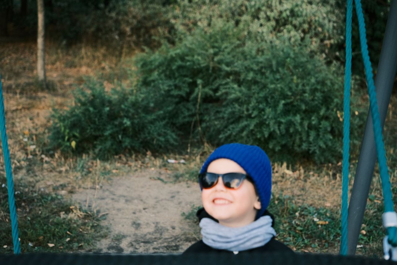 Smiling child in a blue beanie and sunglasses riding an outdoor swing.