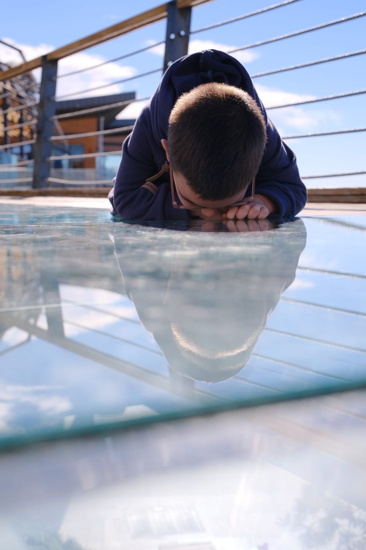 Child lying face-down on a glass viewing platform with a reflected face