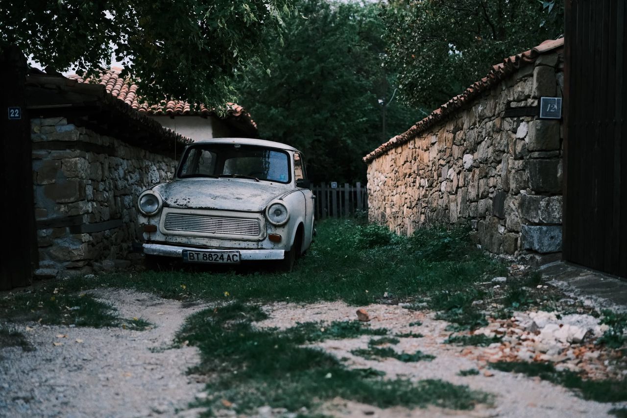 Weathered white car parked between stone walls beneath trees in a Bulgarian village.