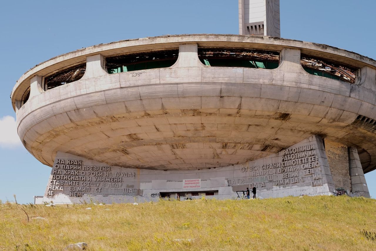 Weathered saucer-shaped Buzludzha monument on a grassy hill, with part of its tower visible behind the circular roof.