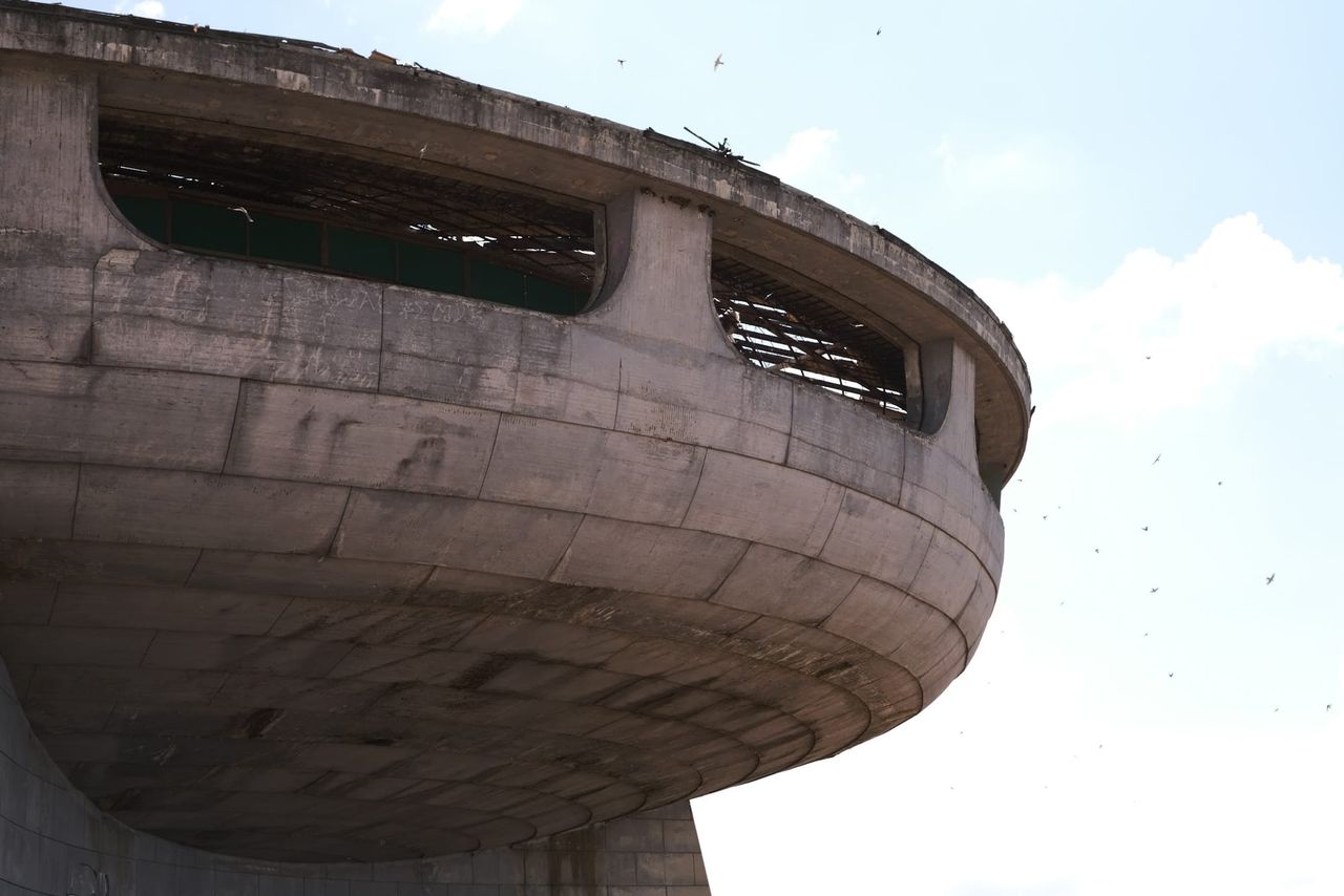 Curved concrete facade and broken window openings of the Buzludzha monument beneath a pale sky.