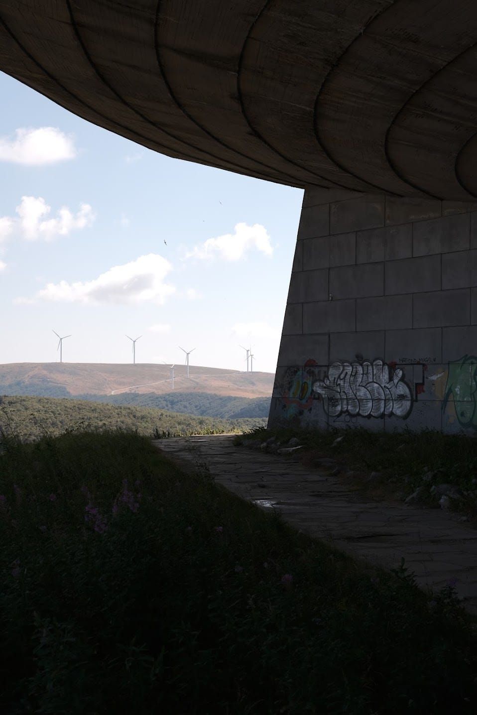 Shaded underside of Buzludzha's curved roof and a graffiti-covered pillar framing distant hills.