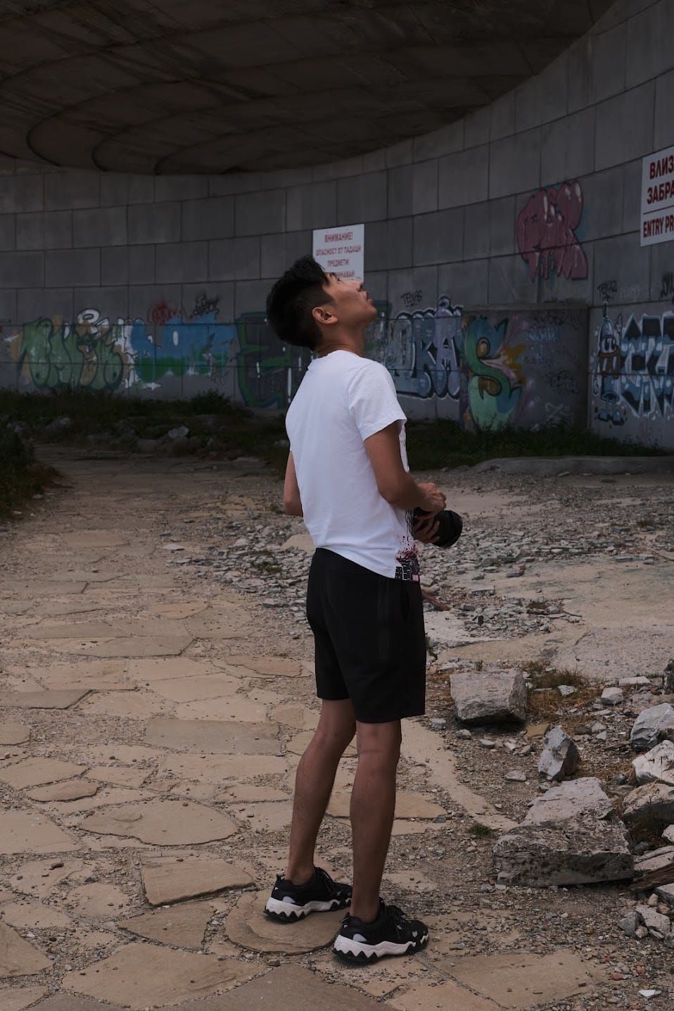 Man in a white shirt and black shorts holding a camera and looking up beneath Buzludzha's graffiti-covered concrete structure.