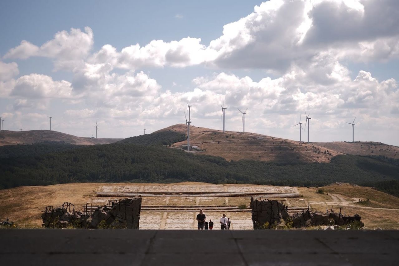 View down Buzludzha's entrance steps across a broad plaza with three visitors, distant hills and wind turbines beneath clouds.