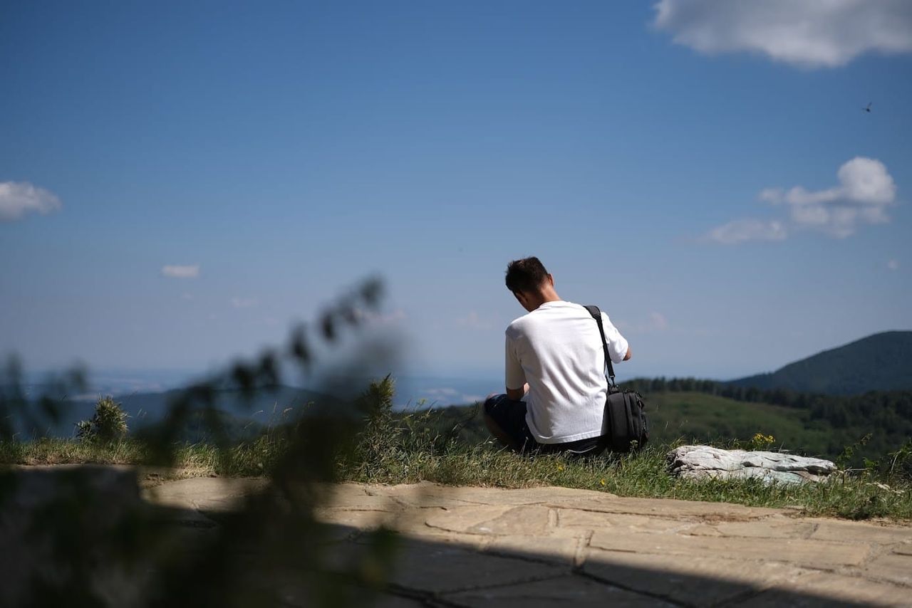 Man in a white T-shirt seated on grass overlooking distant mountains, with soft blurred wildflowers in the foreground near Buzludzha.