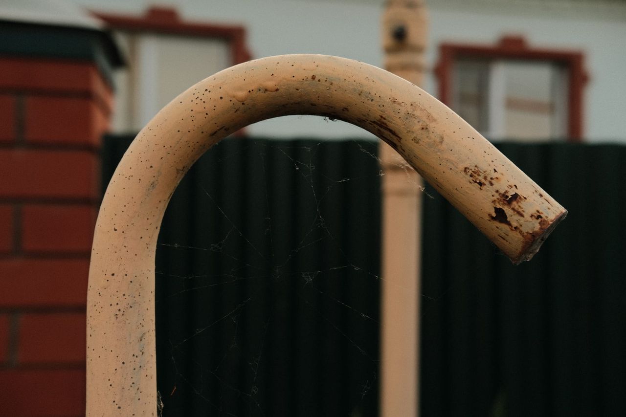 A curved rust-speckled pipe rises in front of a green village fence.