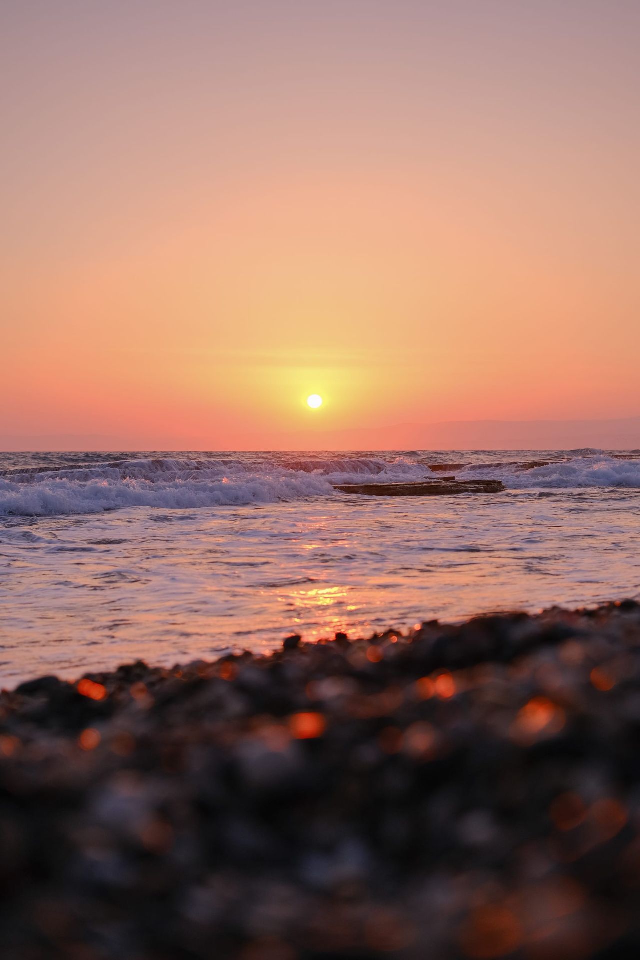 Orange sun setting over gentle sea waves and a dark pebbled shore