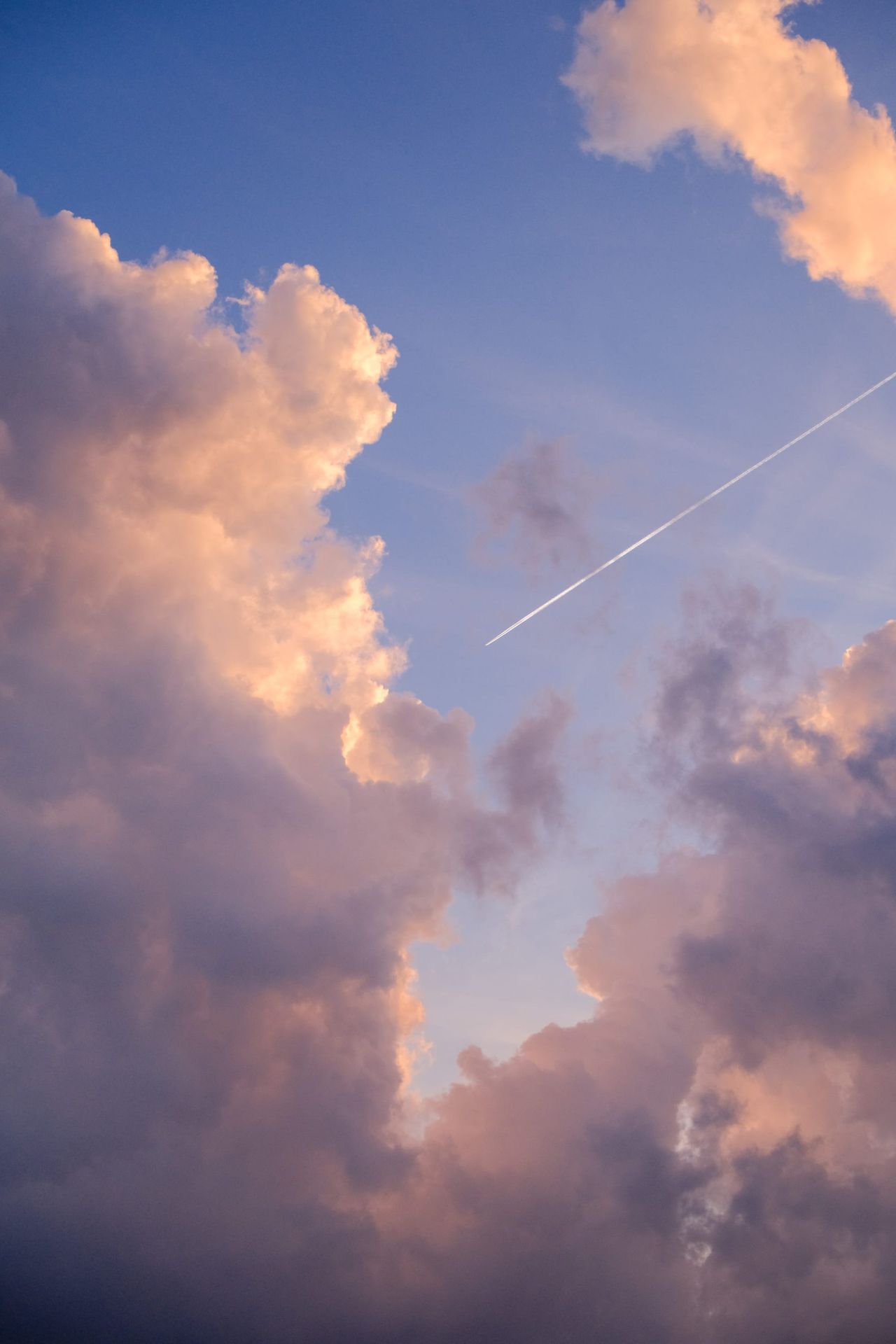 White aircraft trail crossing between peach-lit cumulus clouds