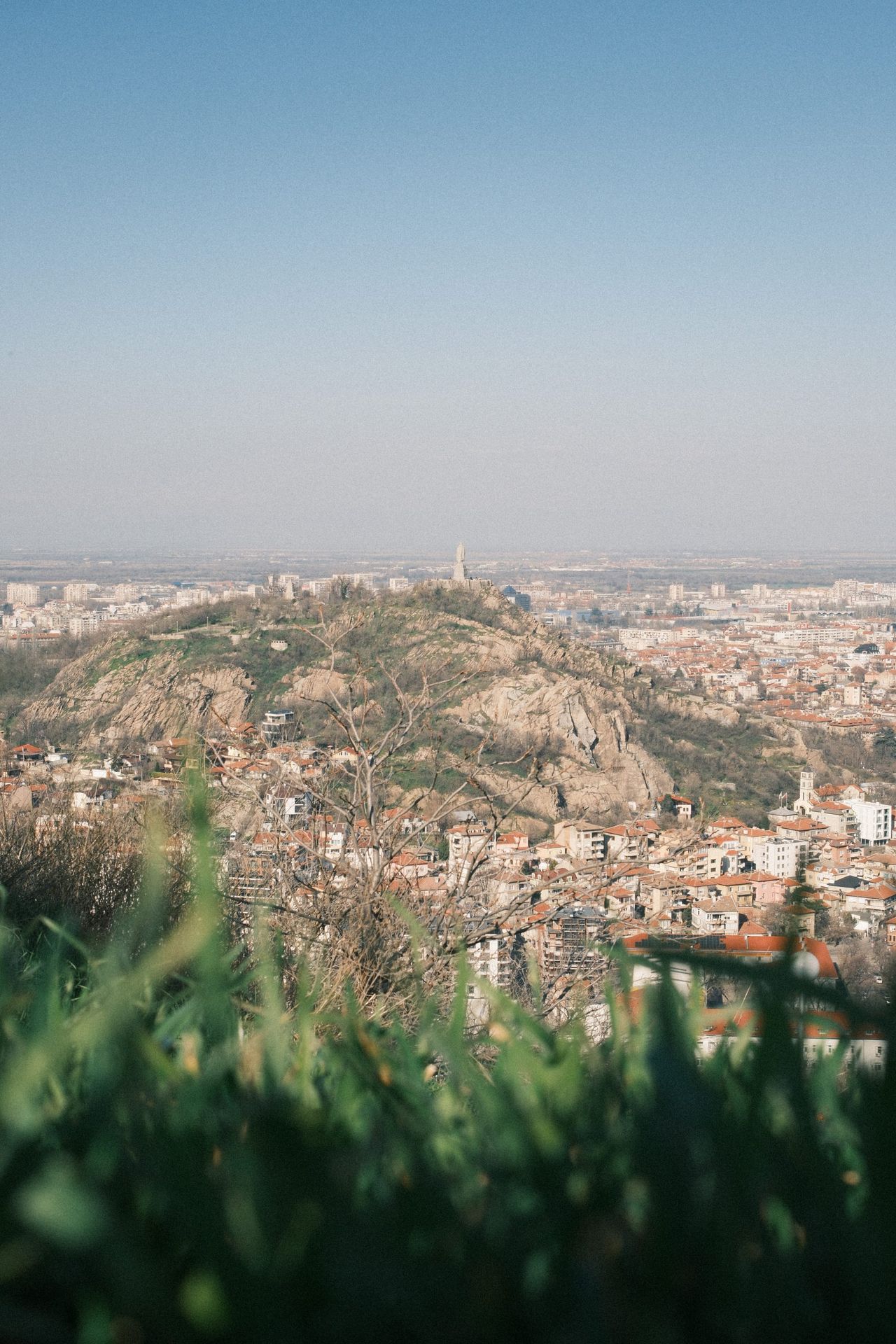 Wide city view from a grassy hillside beneath a hazy pale blue sky