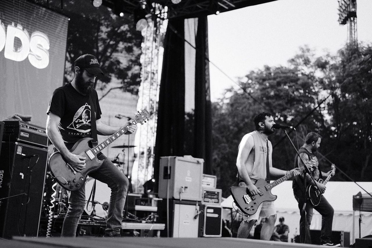 Three members of 1000Mods playing guitars and bass on an outdoor festival stage.