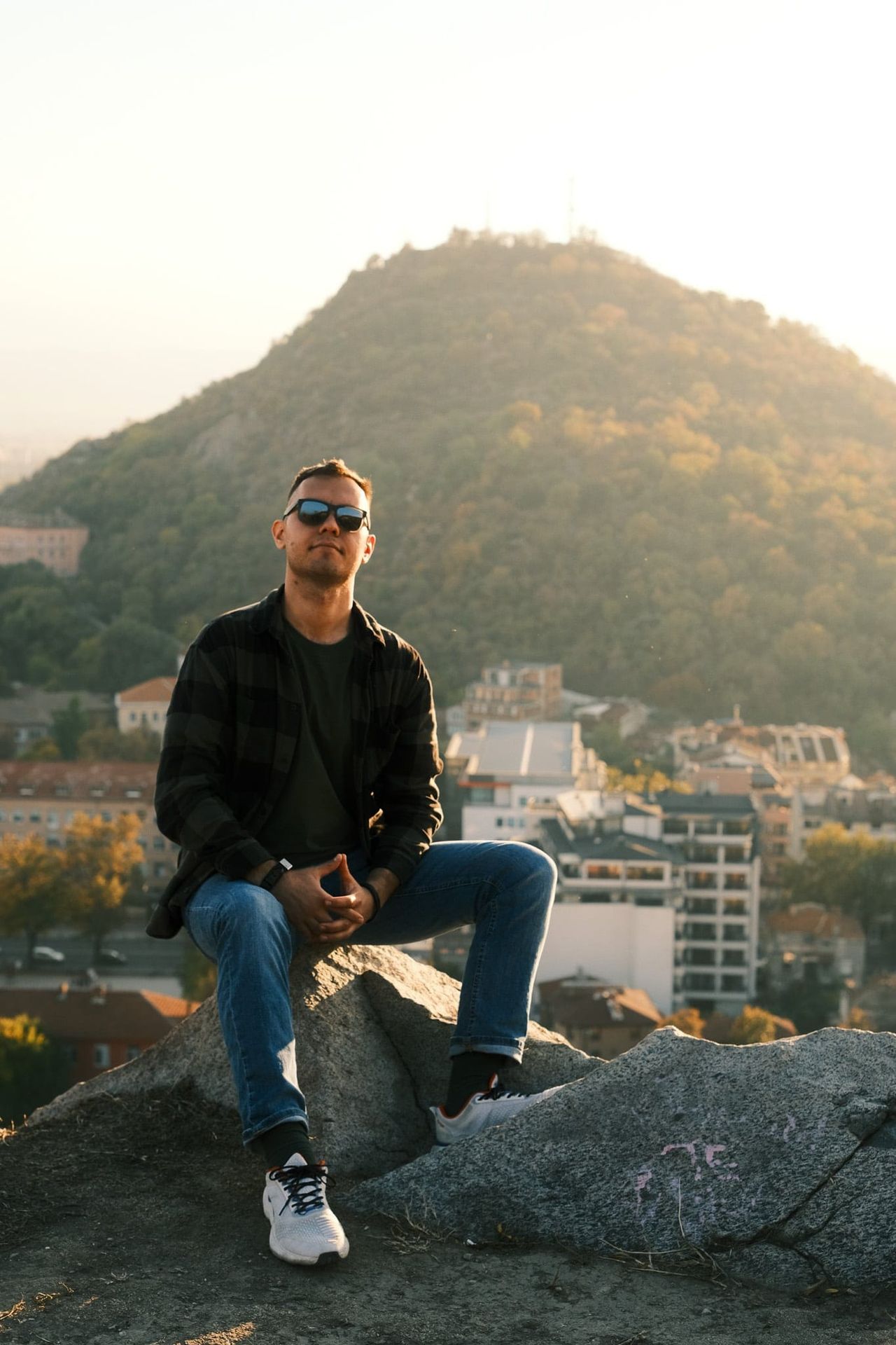 Man in sunglasses sitting on rocks above a city at golden hour.