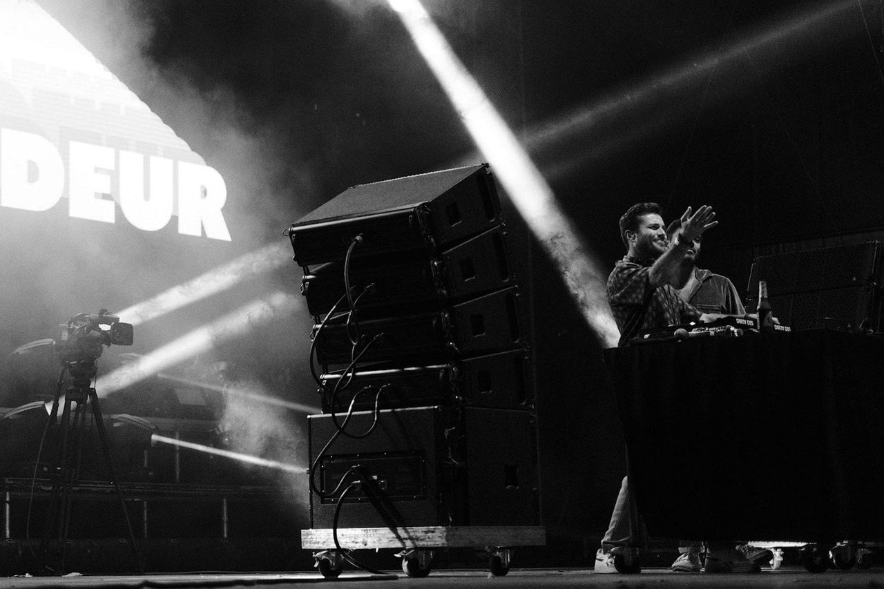 Bon Entendeur performer gesturing behind DJ equipment under crossing stage lights.