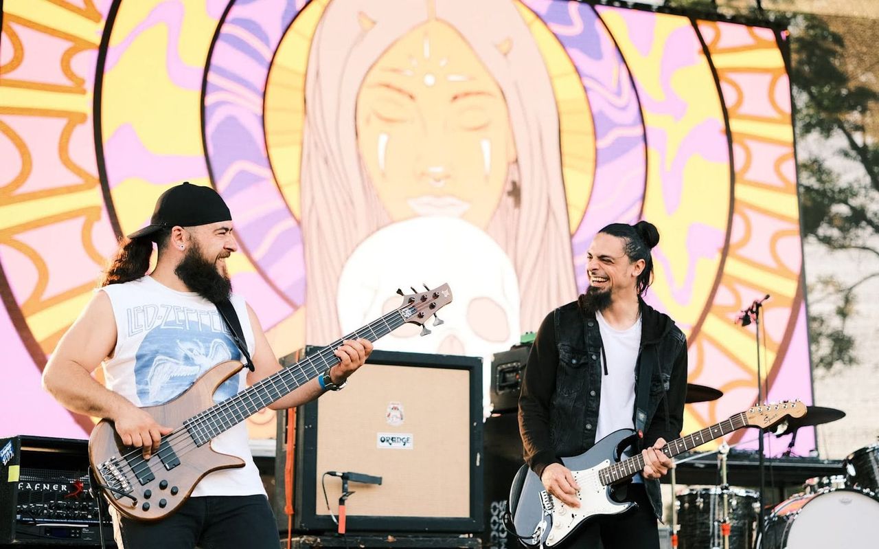 Two Me & My Devil musicians smiling while playing bass and electric guitar onstage.