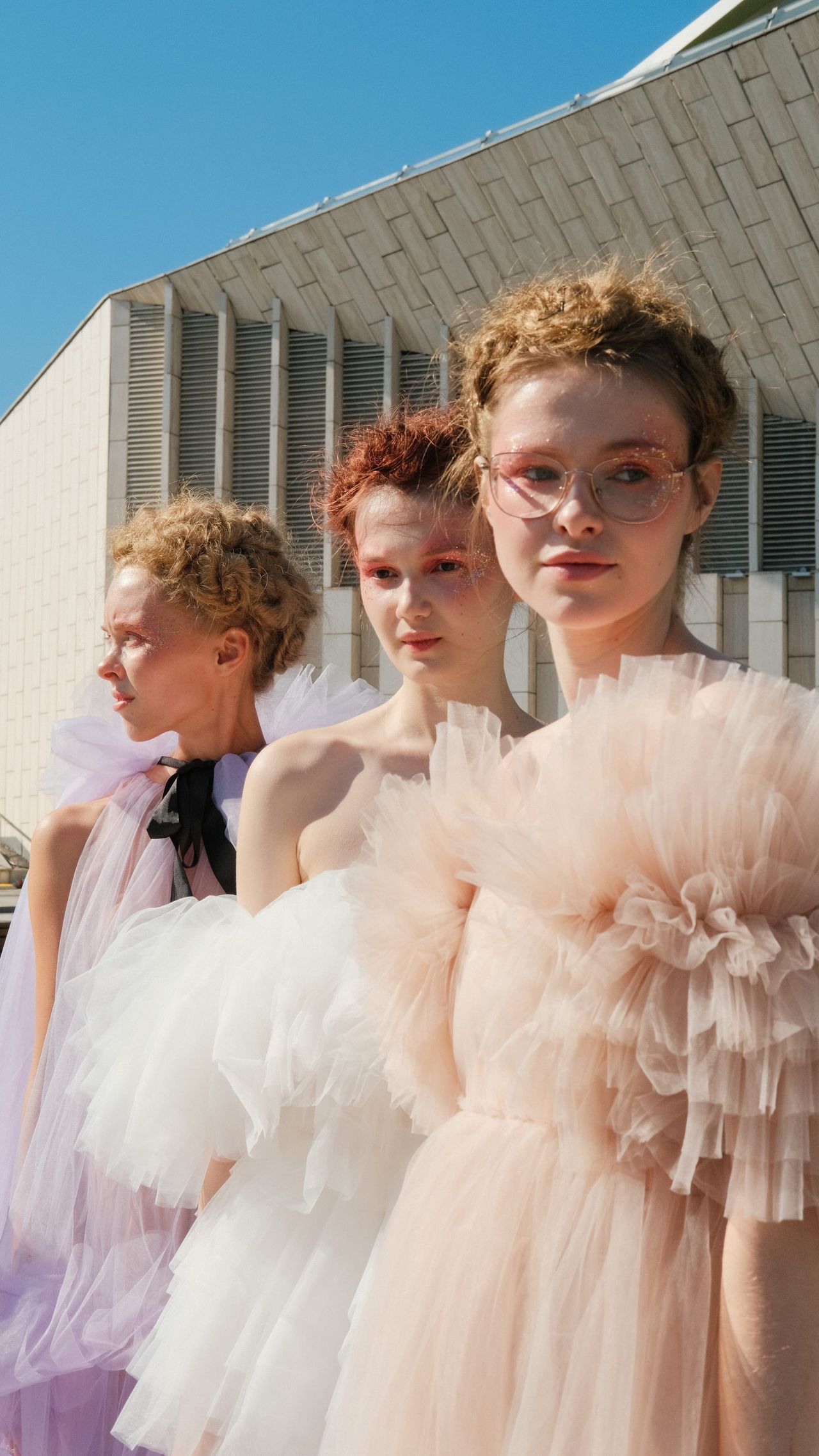 Models in pale pink and white ruffled dresses stand together outside the venue.