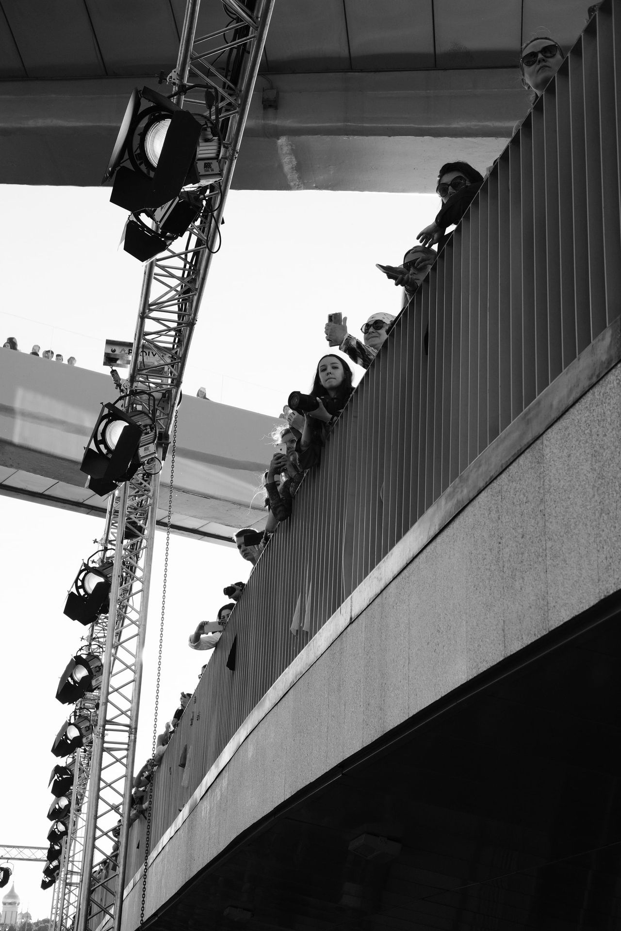 Black-and-white upward view of the venue structure, suspended lights and spectators along an elevated walkway.