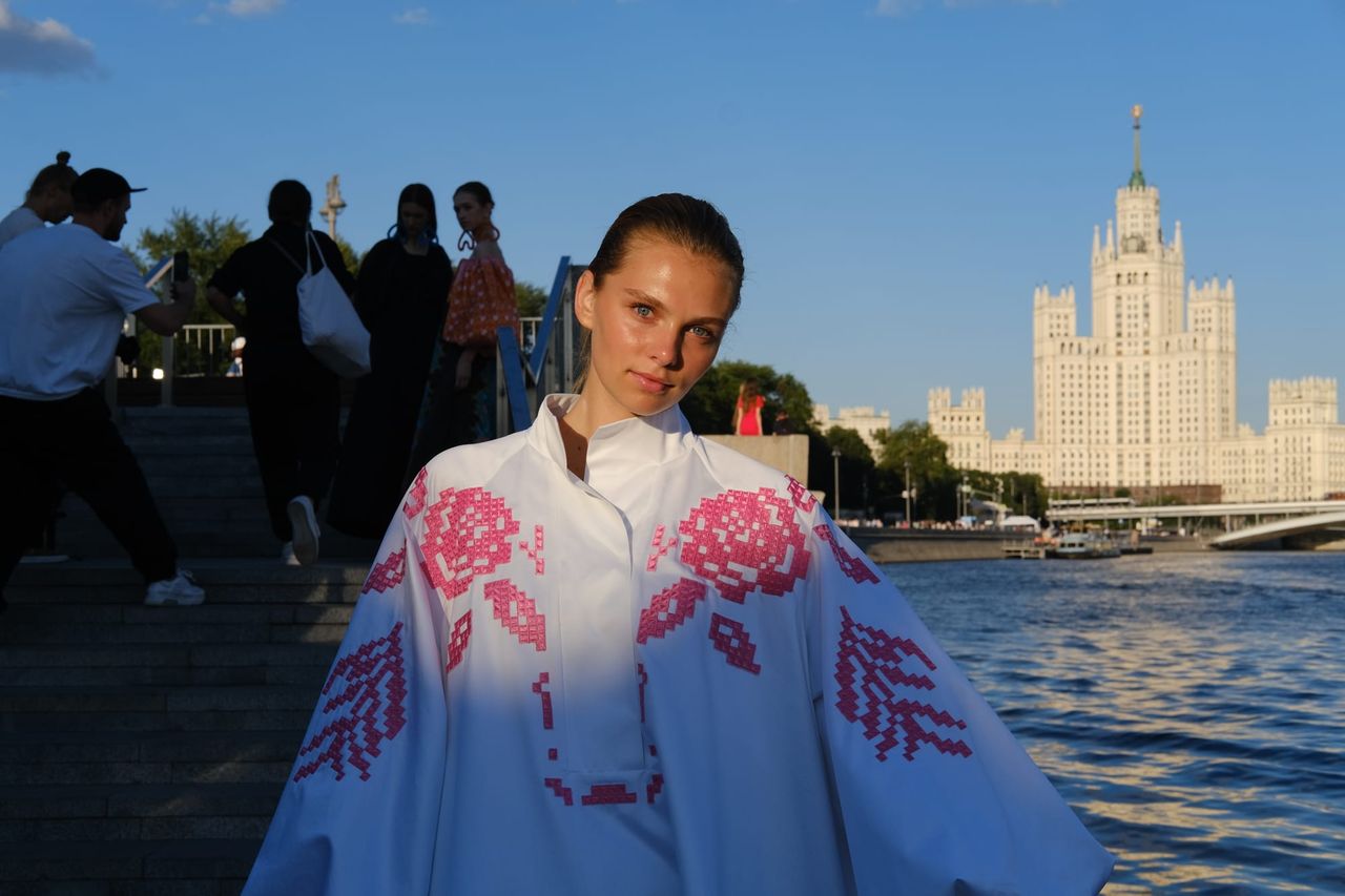 A model wears a white cape-like outfit decorated with pink flowers beside the river, with a tall building across the water.