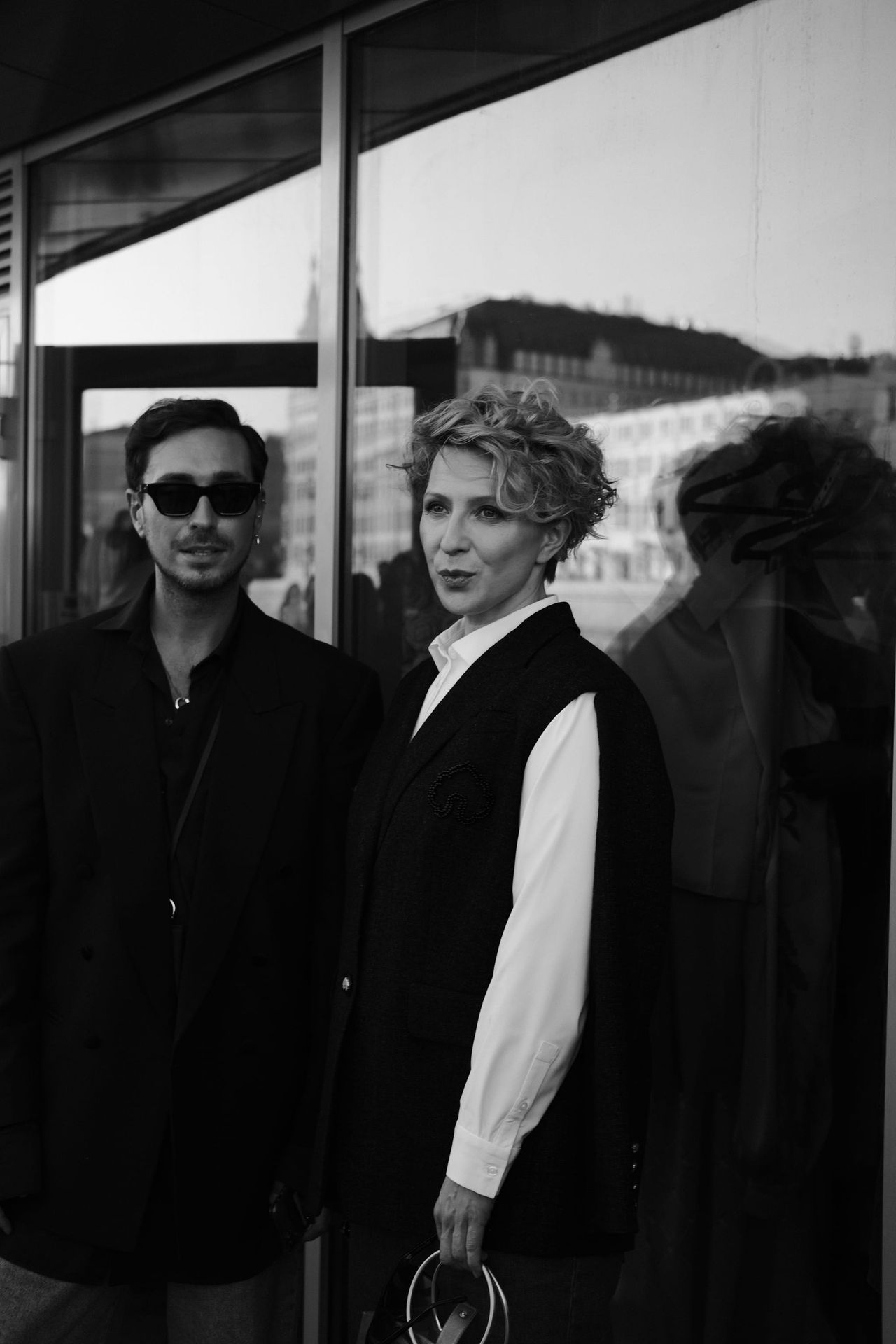 Two guests stand beside the venue’s reflective glass wall in a black-and-white photograph.