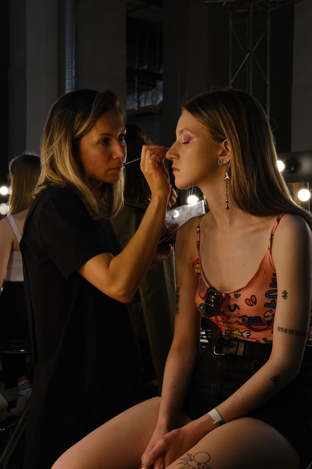 A makeup artist works on a seated model’s face backstage beneath warm lights.