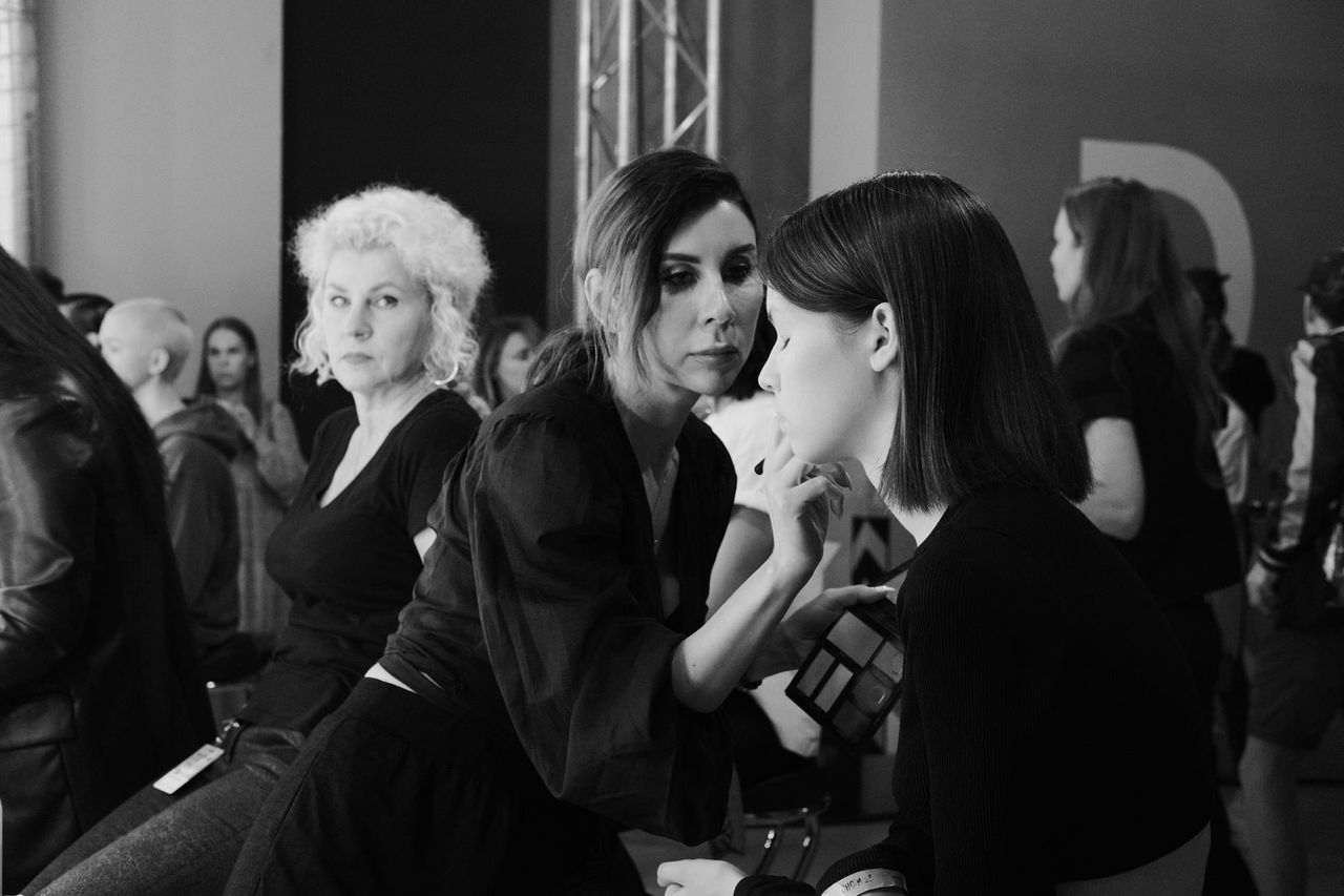 A makeup artist attends to a seated model while other people wait nearby, in a black-and-white backstage photograph.