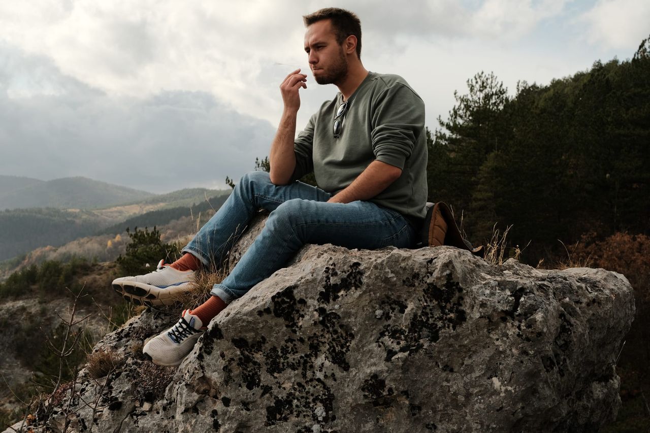 A man sits on a large rock with mountain scenery and clouded sky behind him.