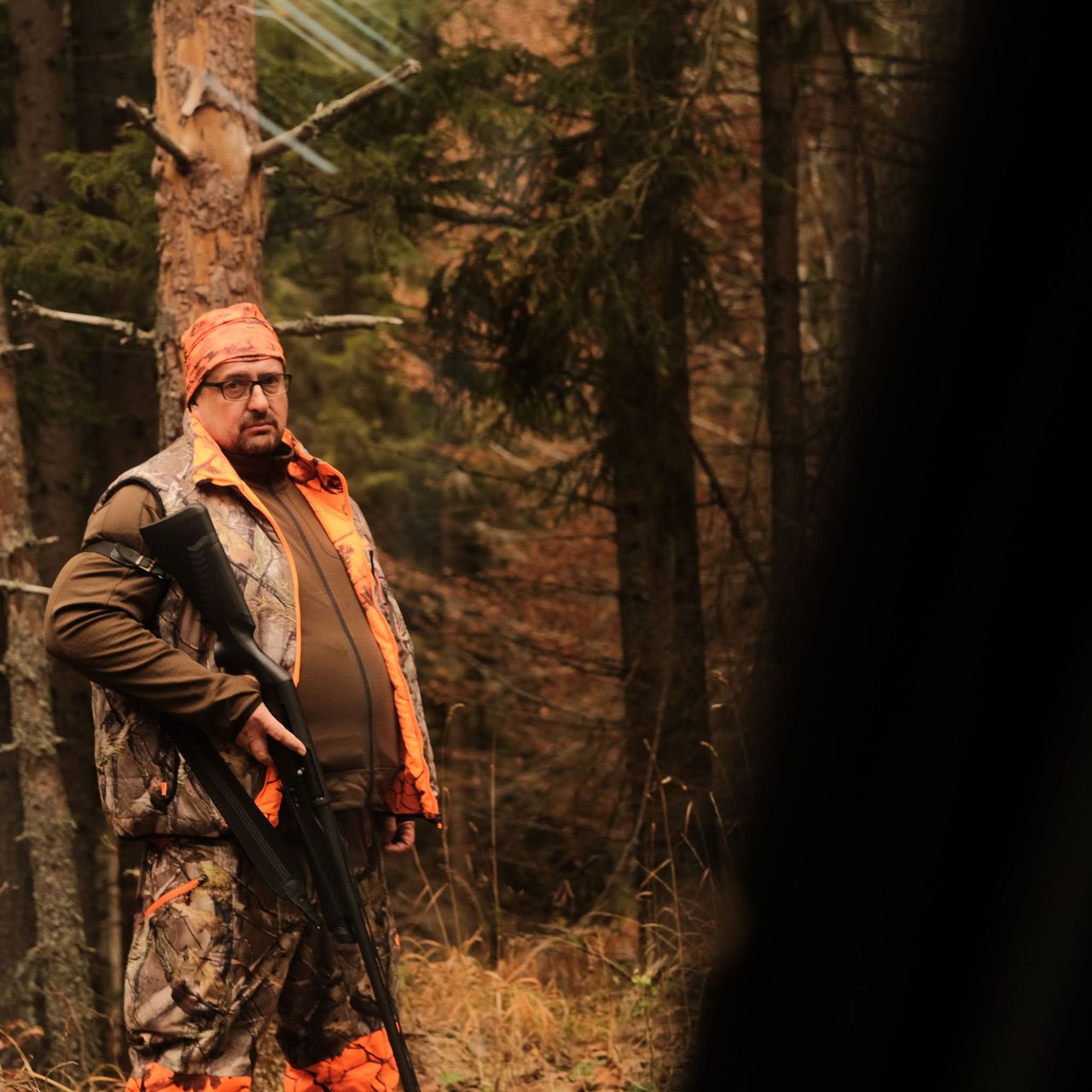 A man in outdoor clothing and orange accents stands among autumn woodland trees.