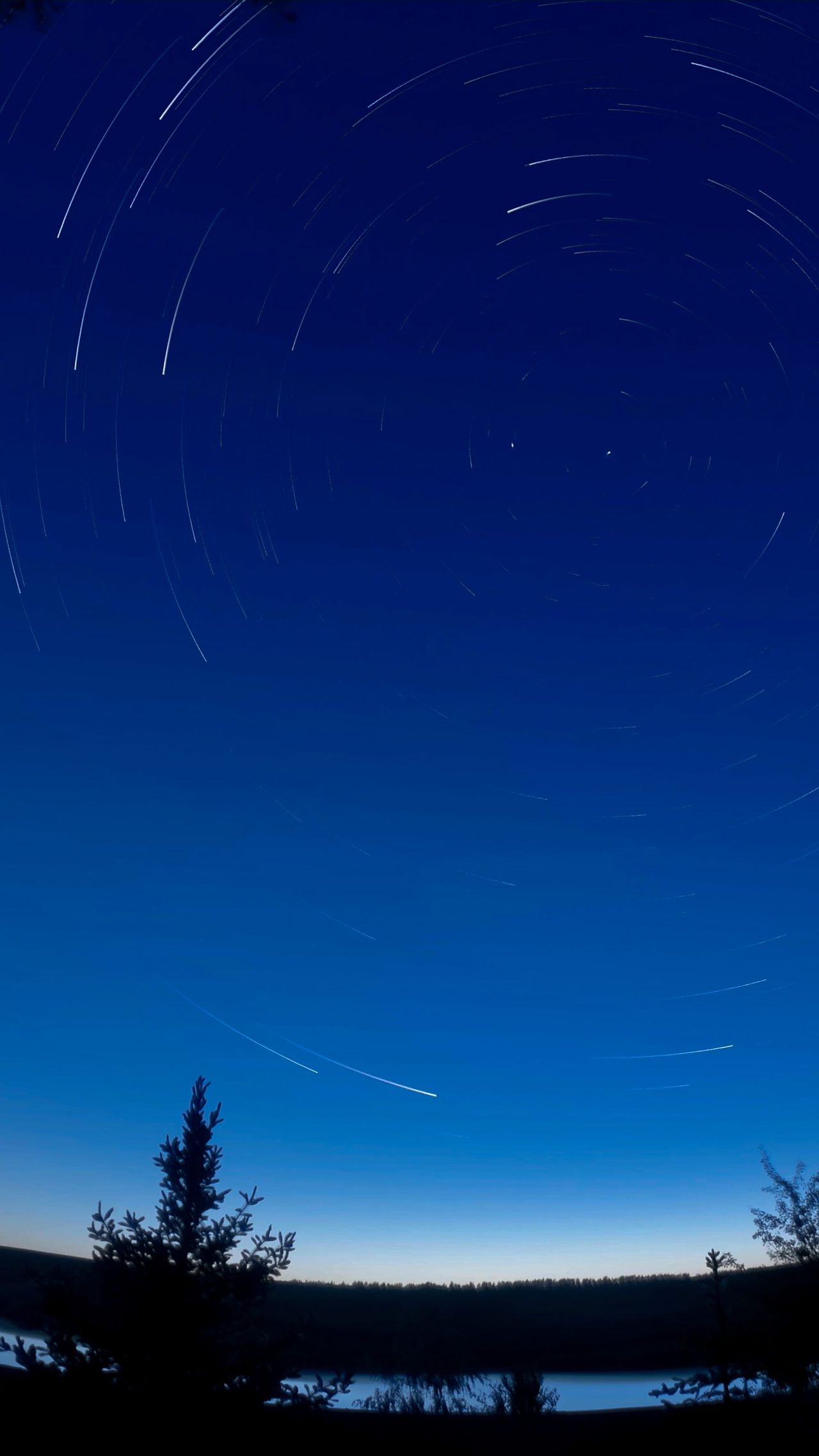Circular star trails above a dark lakeside horizon in a deep blue night sky