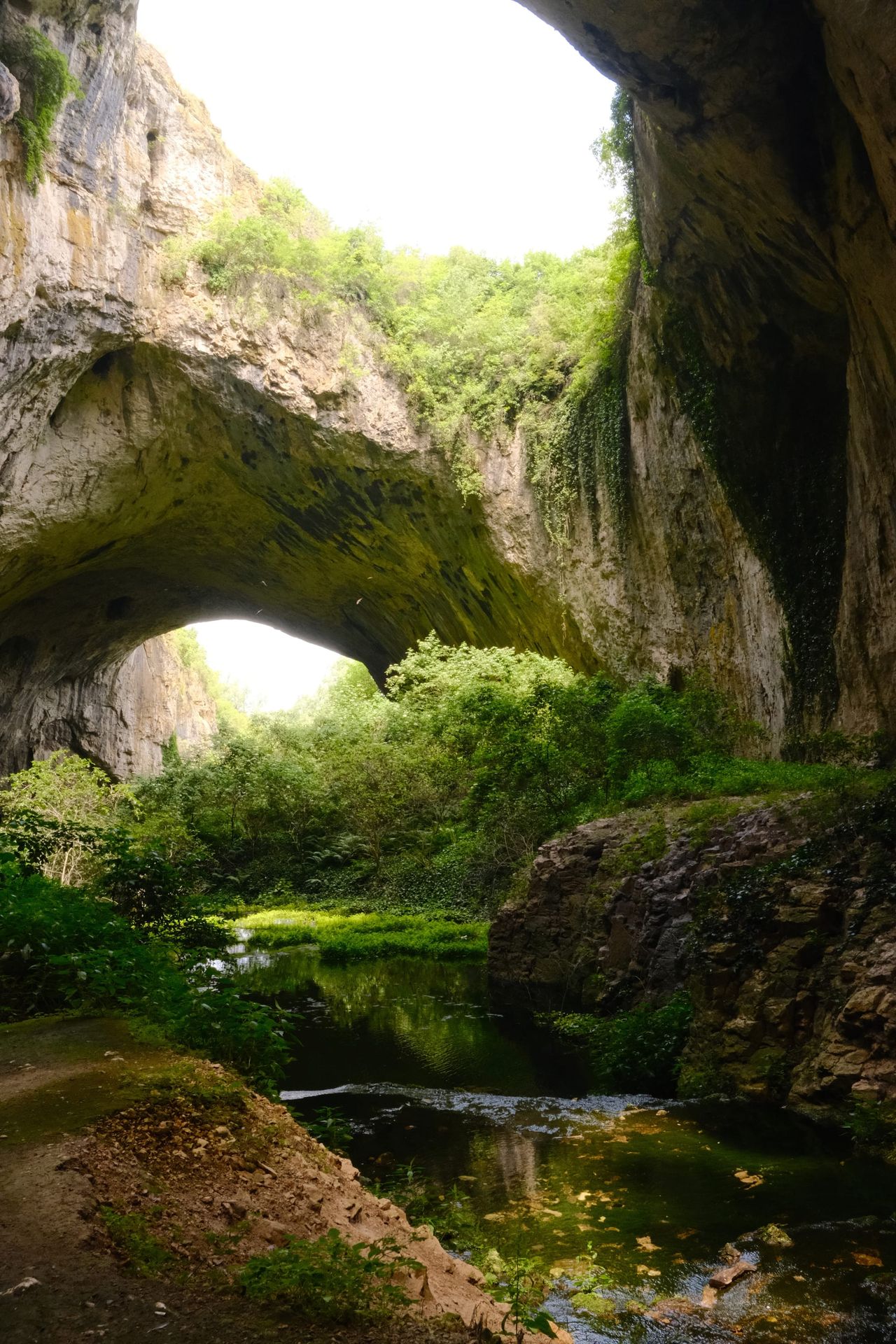 Tall Devetashka arches above a stream and green cave floor