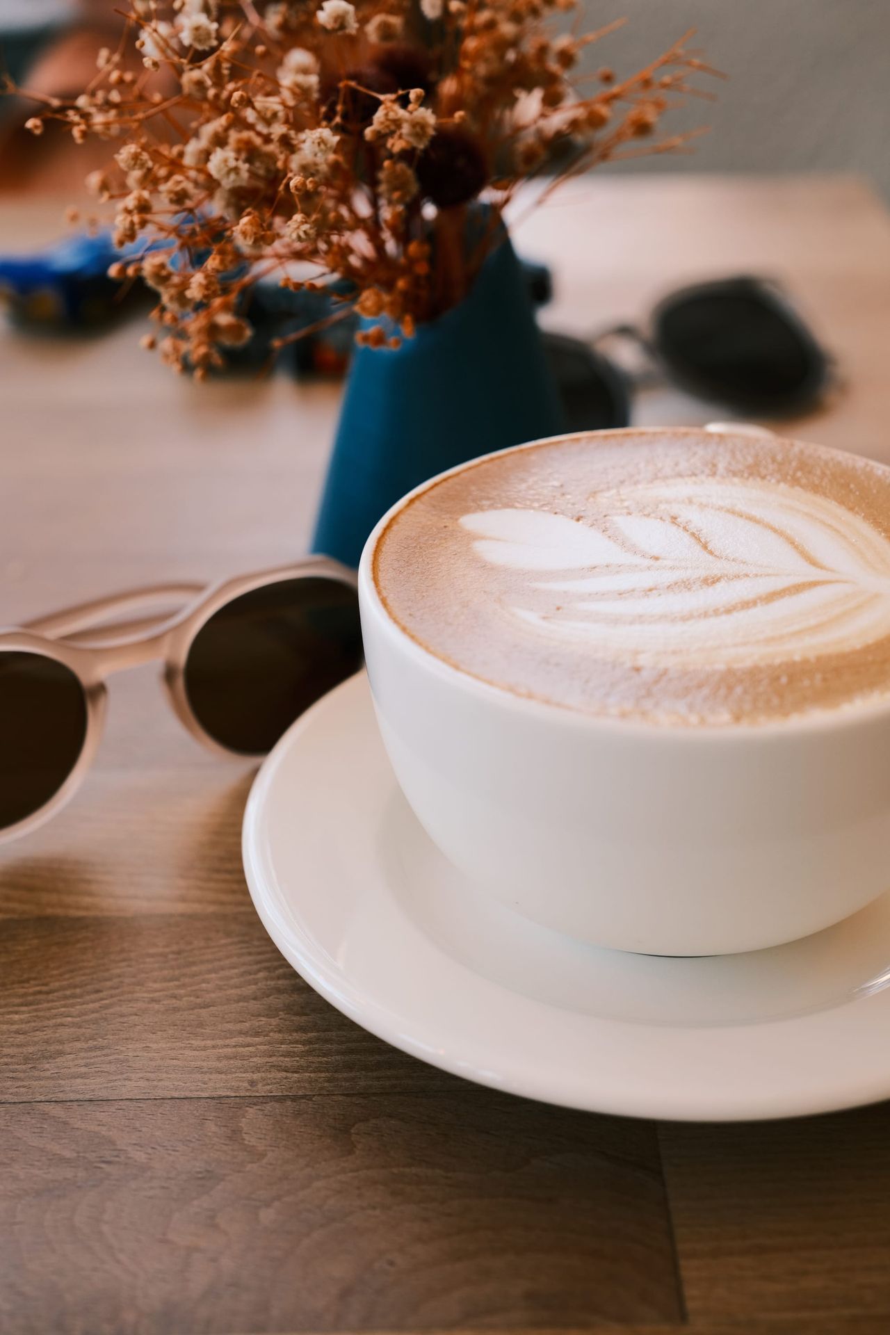 Foamy coffee in a white cup beside sunglasses and dried flowers