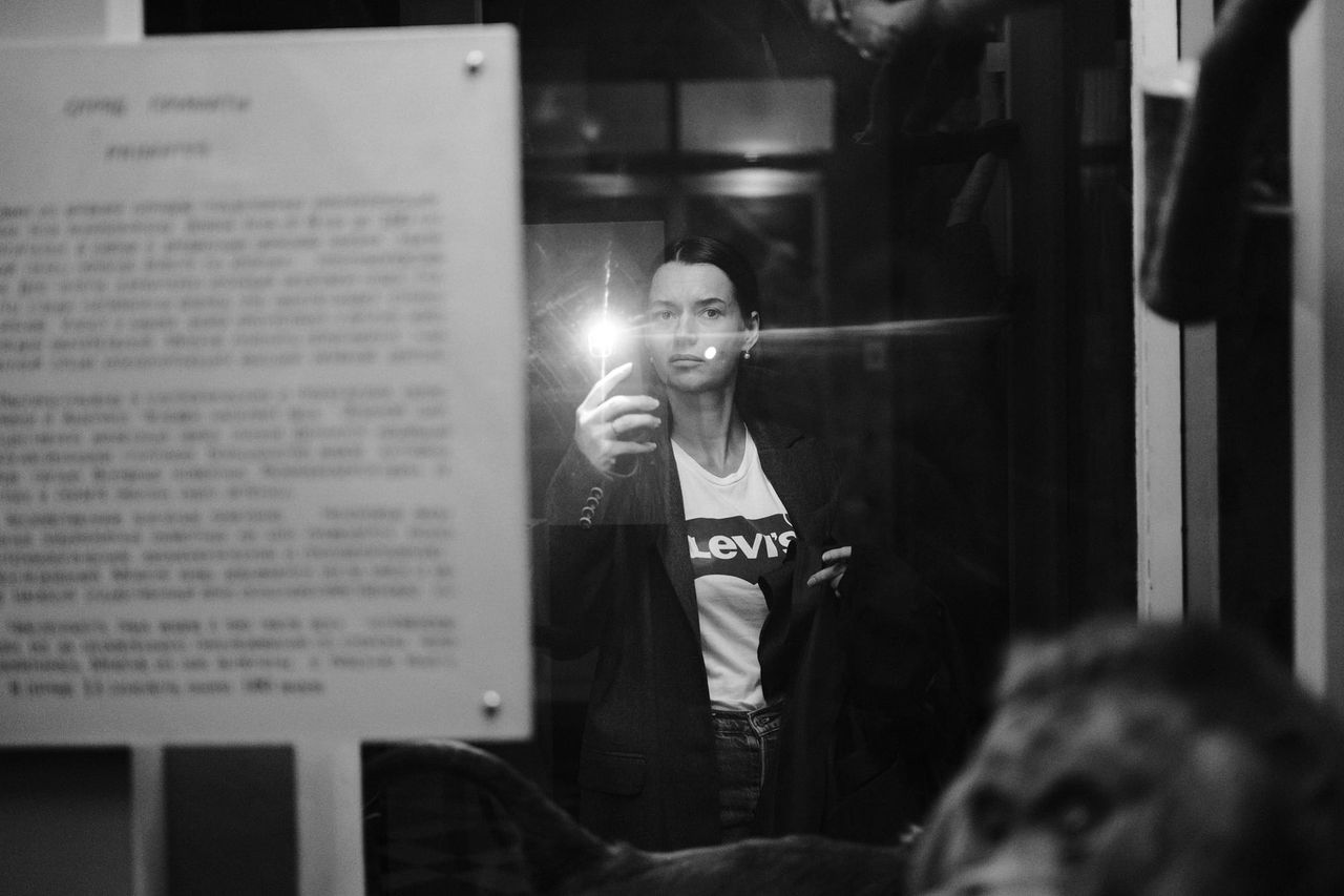 Black-and-white photograph of a visitor holding a lit phone beside a museum information panel.