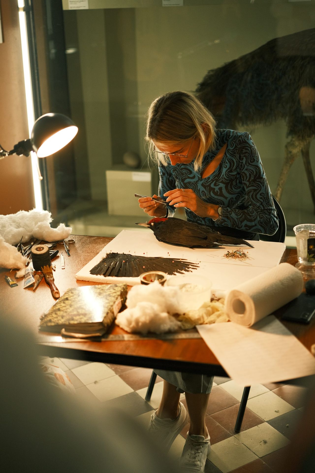 A woman leans over a specimen at a workbench beneath a bright desk lamp.