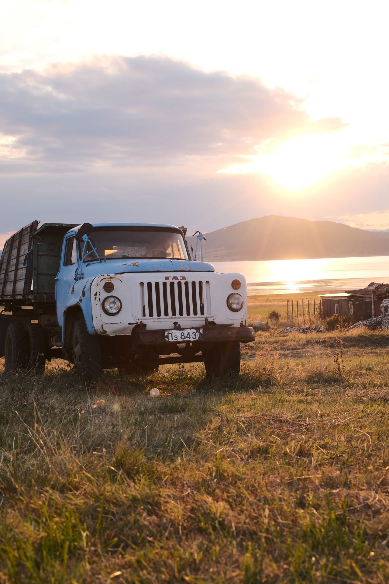 Old blue and white truck parked in grass beside Batak Lake, backlit by the low evening sun.