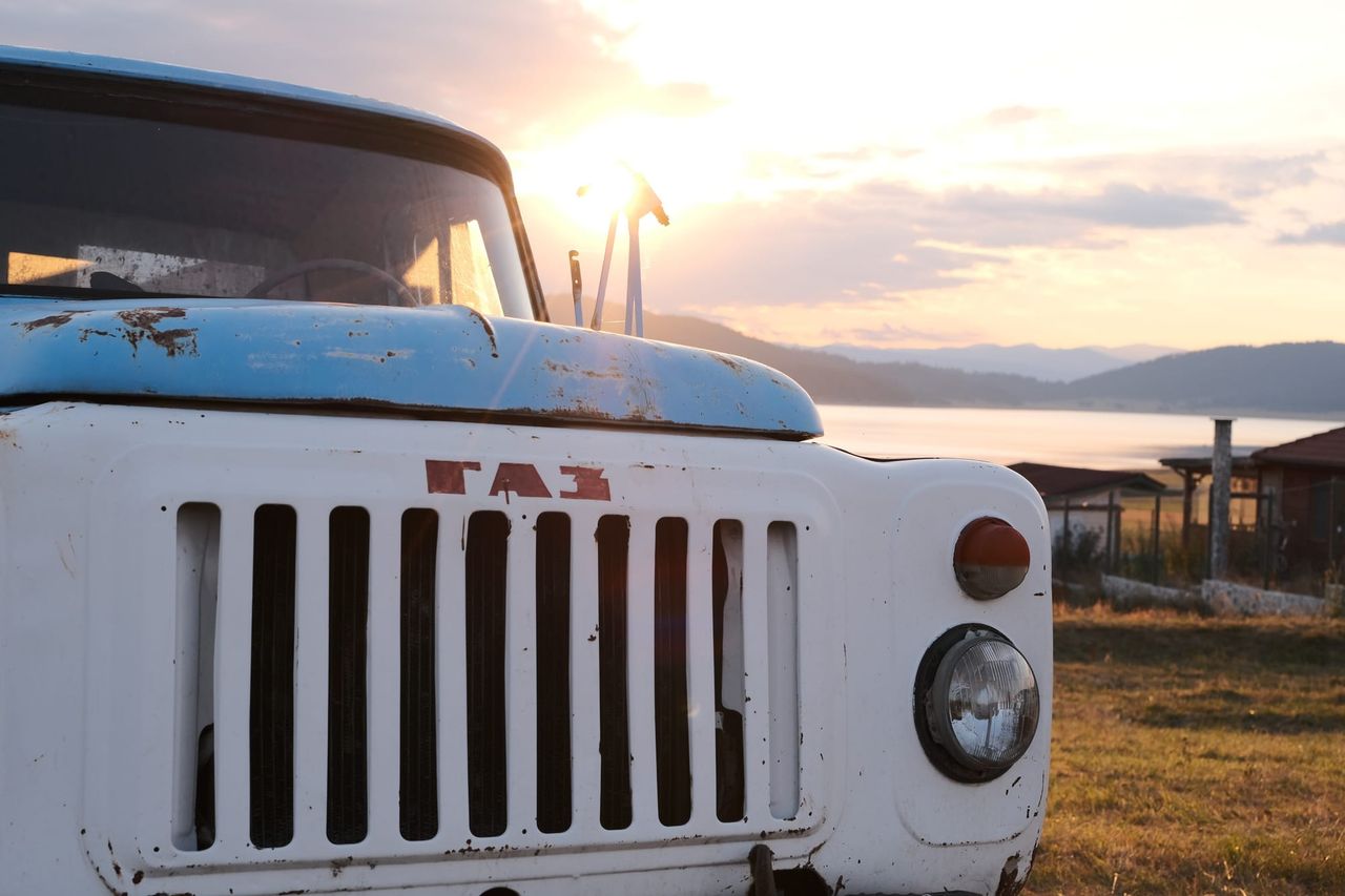 Round headlights, white grille and a weathered blue bonnet on an old truck beside Batak Lake at sunset.