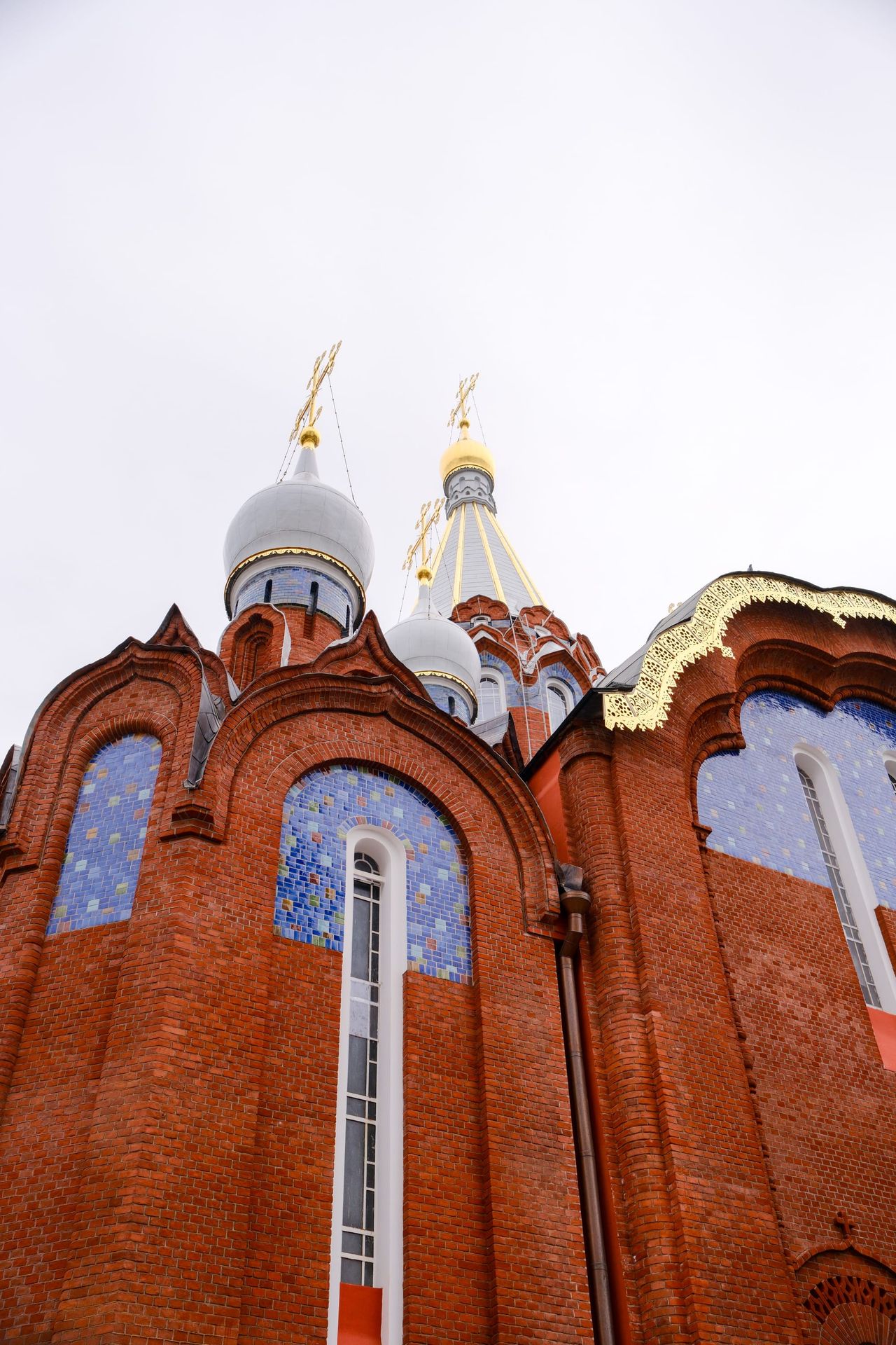 Low-angle view of a red-brick Orthodox church with blue mosaic-tiled arches, silver onion domes, a gilded dome and gold crosses against a pale sky