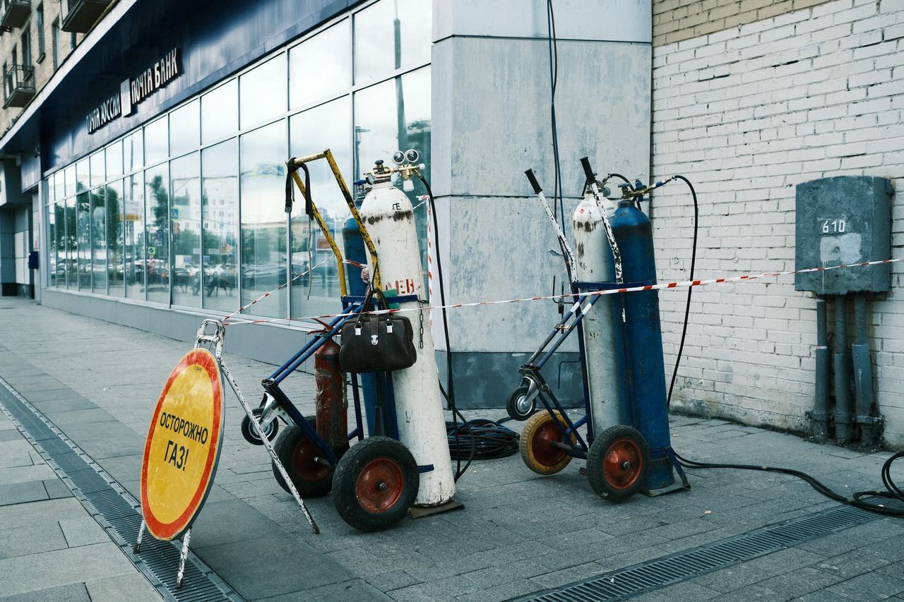 Gas cylinders on wheeled trolleys stand beside a white brick wall and a yellow warning sign on a Moscow pavement.