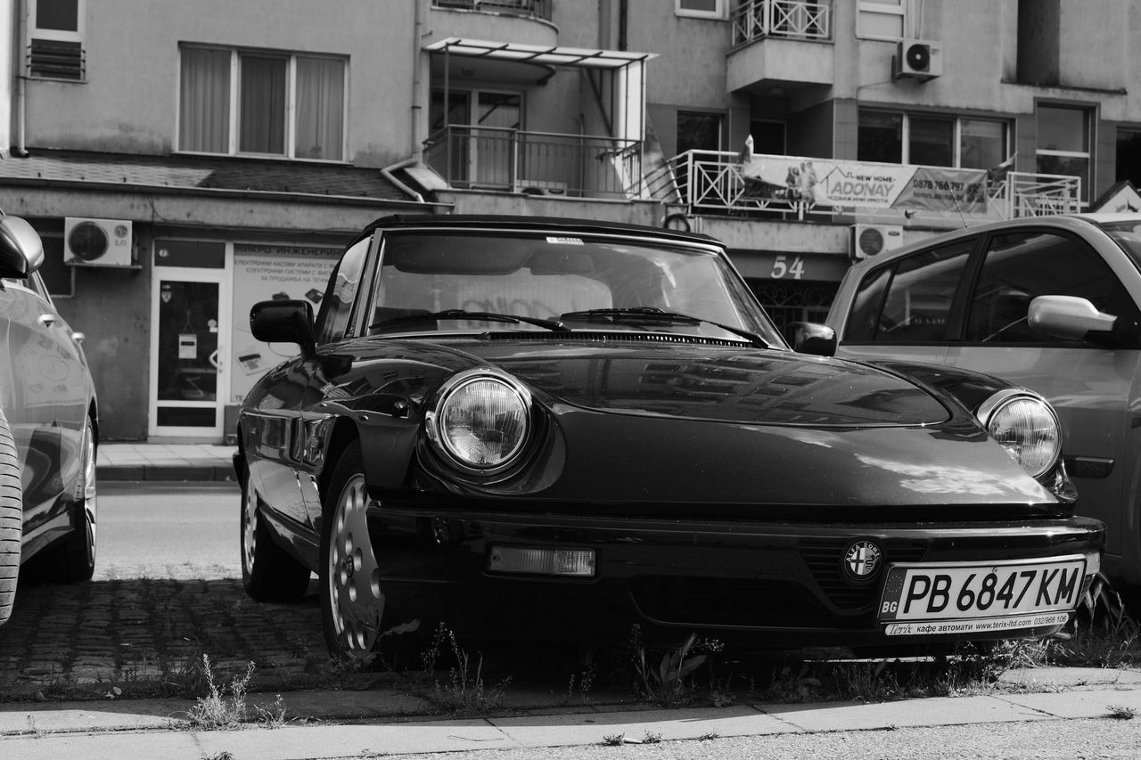 Black and white photograph of a parked sports car with round headlights in front of apartment buildings in Plovdiv.