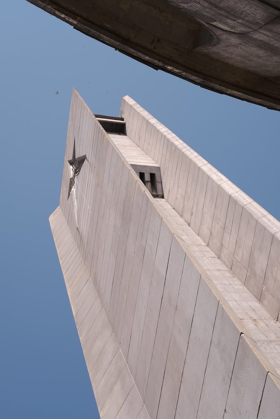 Angular concrete tower of Buzludzha viewed from below, framed by a curved roof edge against a blue sky.