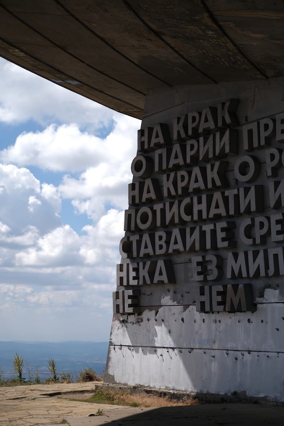 Lettering on a Buzludzha wall beneath a dark concrete overhang, with bright clouds visible to one side.