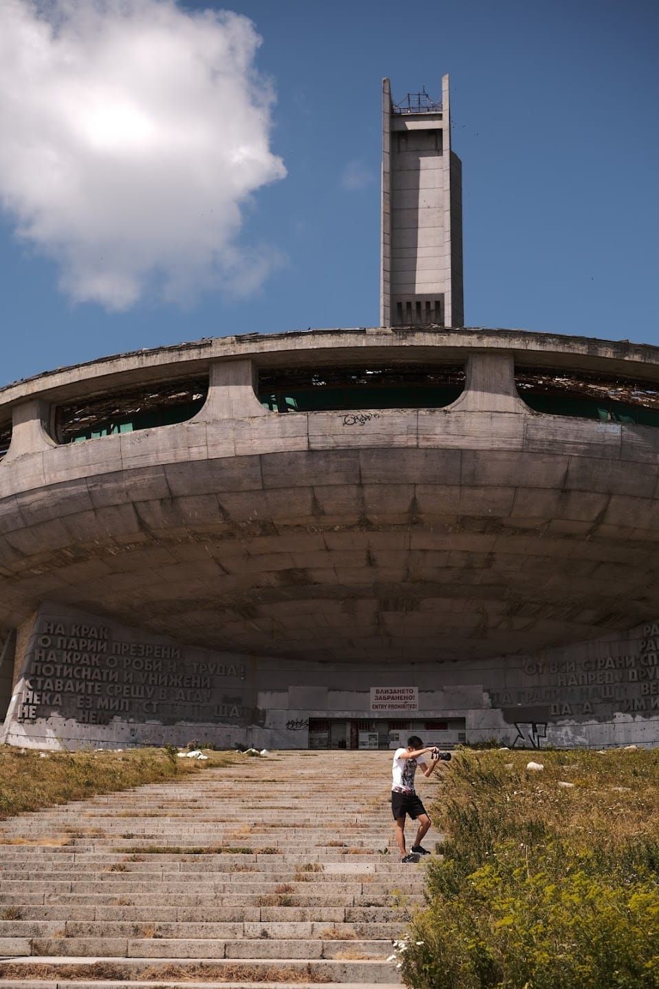 Buzludzha's circular concrete monument, tall tower and broad steps, with a small walking figure showing the scale.
