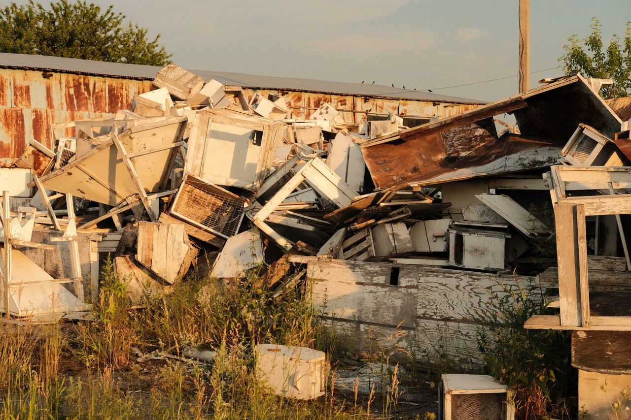 A large heap of weathered wooden boxes and boards lies beside a long farm building.