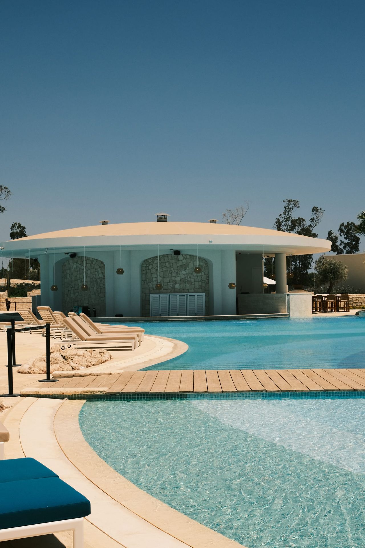 Piscine turquoise incurvée avec passerelle en bois, rangées de transats blancs et un bar rond blanc sous un ciel dégagé.