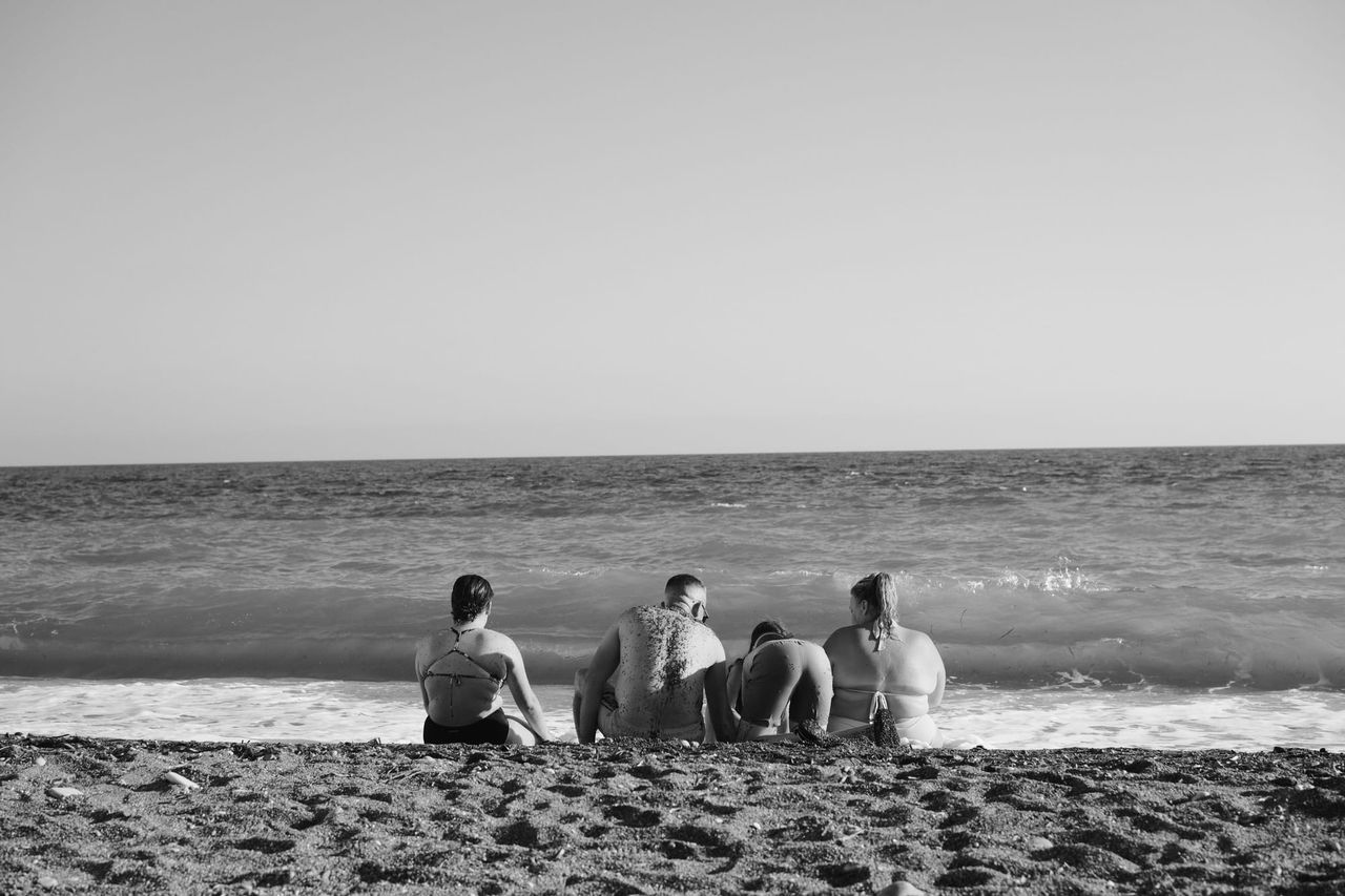 Black-and-white photo of four people seen from behind, sitting close together on a sandy beach and looking at the waves.