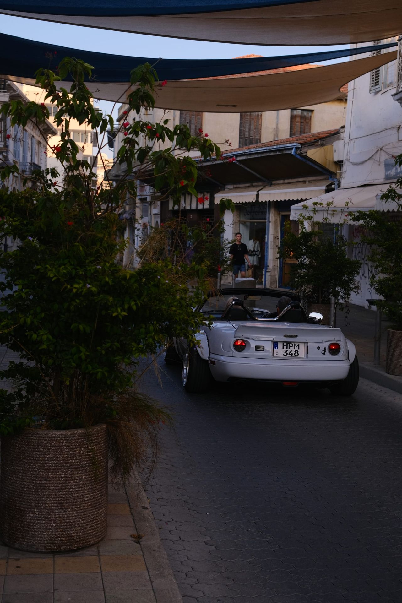 A white classic two-seater convertible parked on a narrow cobbled street under shade sails, beside a large potted tree and cafe awnings.
