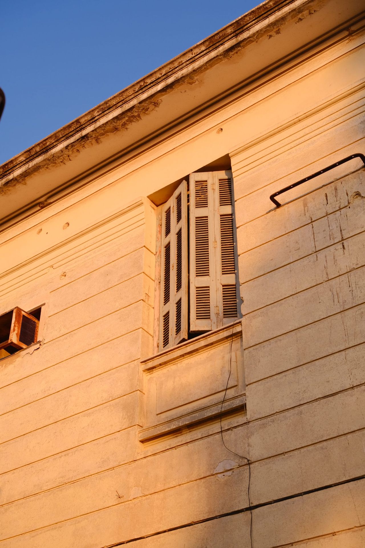 Half-open wooden louvred shutters on a tall window in a cream-coloured plaster wall glowing in warm late sunlight under a blue sky.