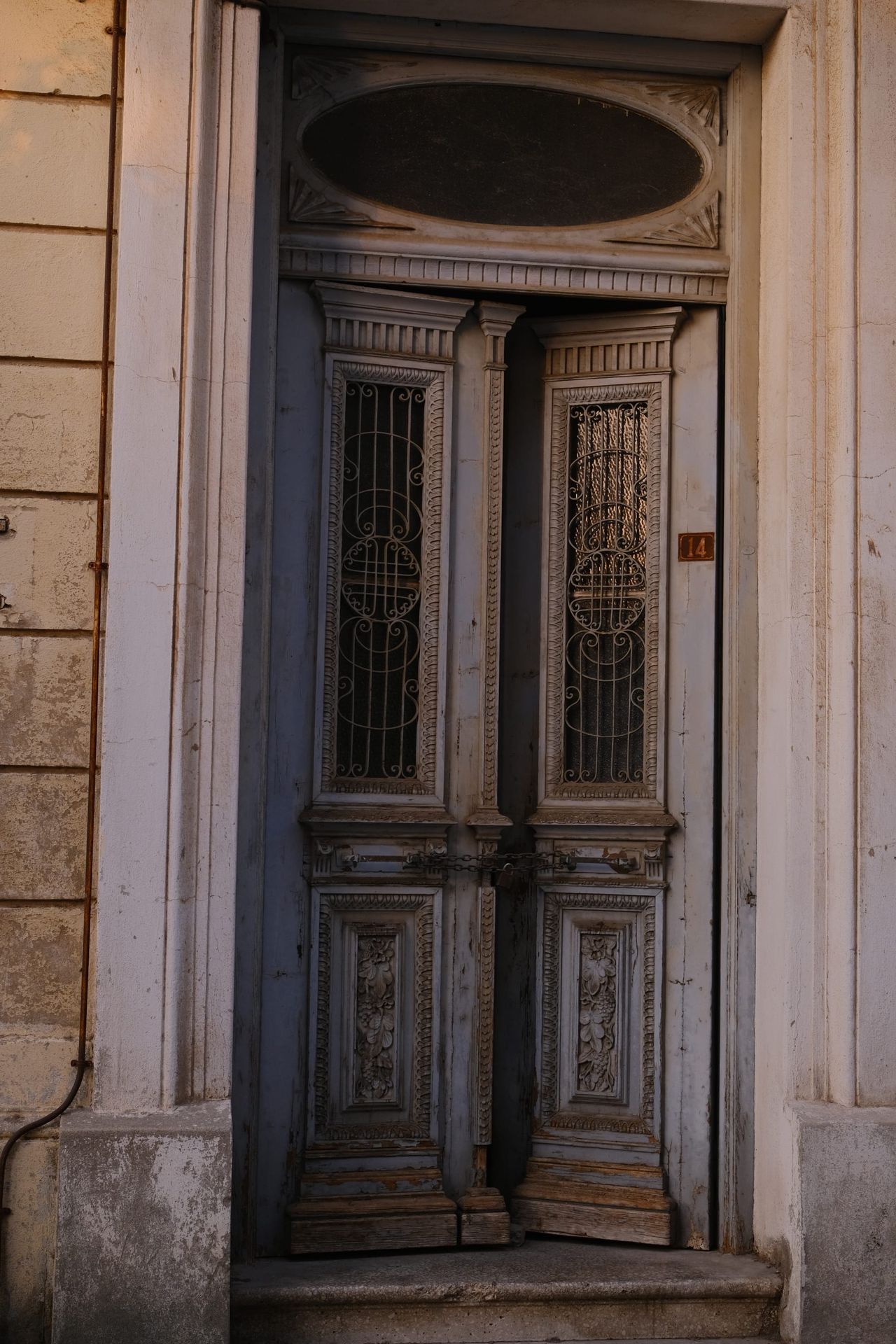 Une haute porte double en bois bleu pâle aux panneaux sculptés, avec grilles en fer forgé et imposte ovale, dans un mur de pierre au-dessus d'une marche usée.