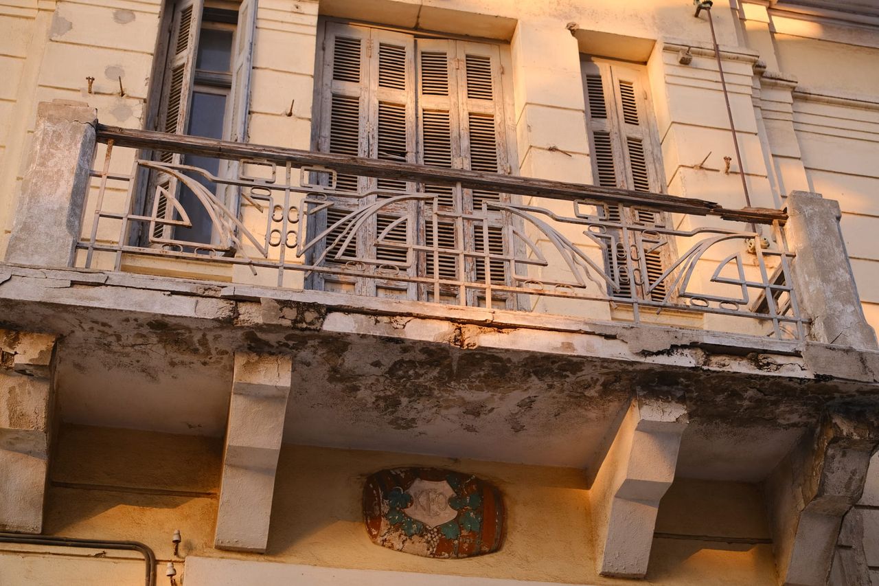 A crumbling stone balcony with a curving wrought-iron railing in front of tall wooden louvred shutters on a cream facade lit by warm evening sun.