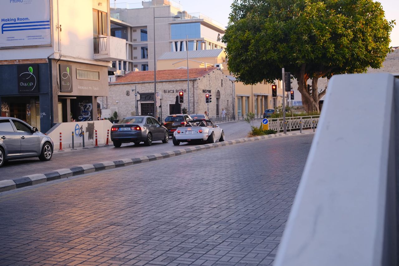 A wide paved street curving past parked cars, a white classic convertible, traffic lights and a stone building with a tiled roof, under a large tree in warm evening light.