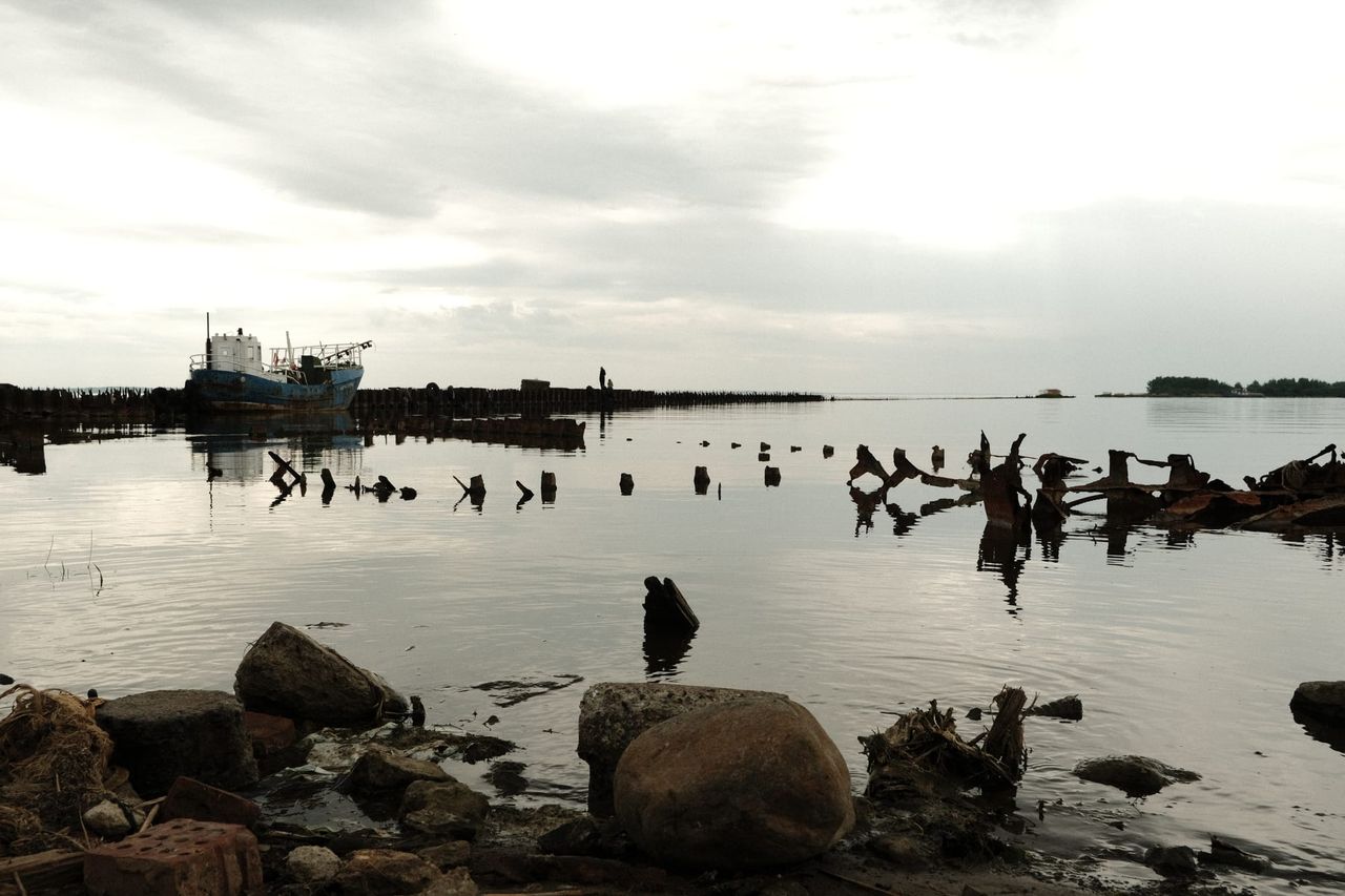 A still grey bay with rotting wooden pier posts, rocks on the shore and a moored ship in the distance.