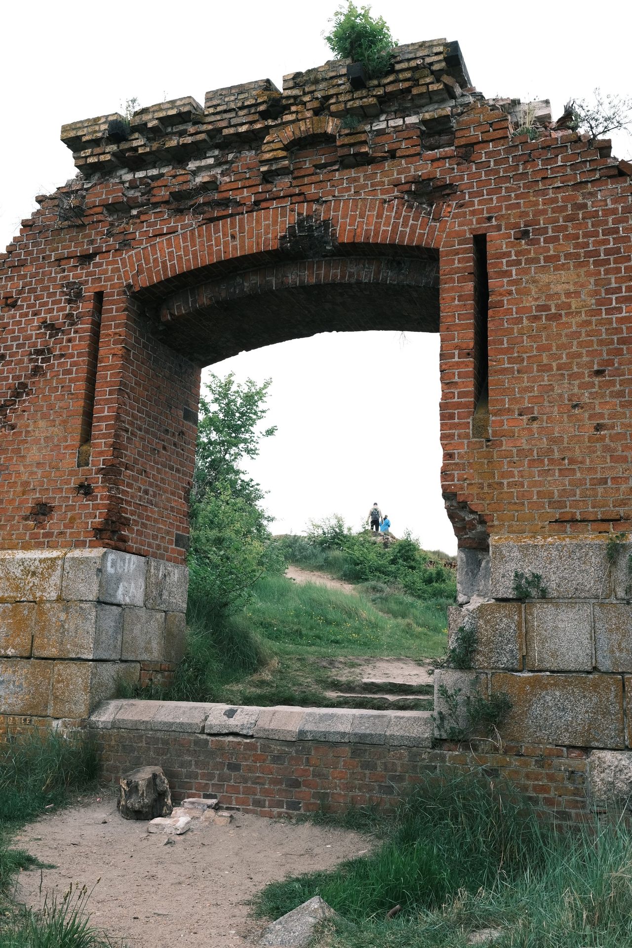 Un arco de ladrillo rojo en ruinas sobre bloques de piedra enmarca una ladera con hierba y dos pequeñas figuras al fondo.