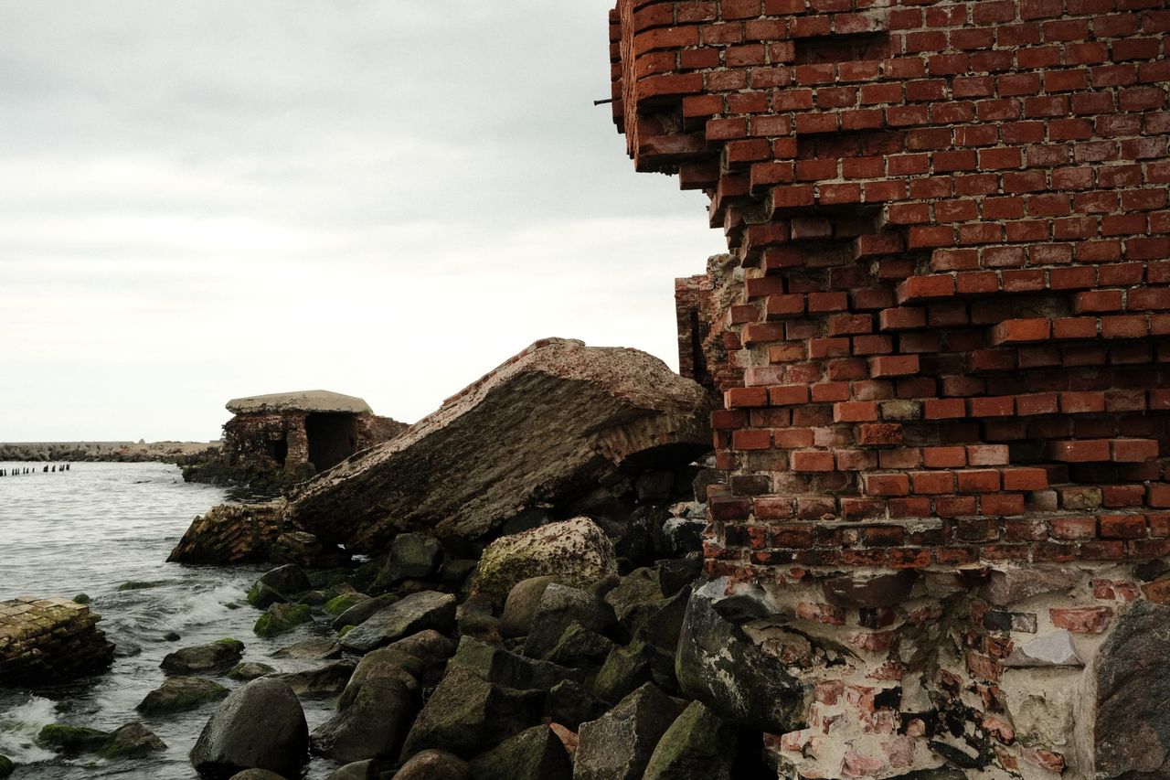 A crumbling red-brick wall beside fallen concrete slabs and mossy boulders at the water's edge, with a small ruined bunker beyond.